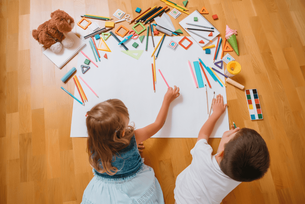 Two young children are lying on the floor and drawing on a large white sheet of paper with colored pencils. They are surrounded by various art supplies, including markers, rulers, geometric shapes, scissors, notebooks, and a watercolor paint set. A stuffed brown dog toy is also visible in the upper left corner. The scene takes place on a wooden floor, and the children appear to be deeply engaged in their creative activity.